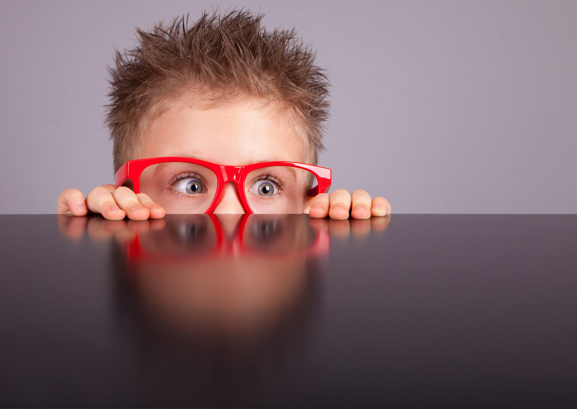 Anxious young boy hiding behind table with with red glasses