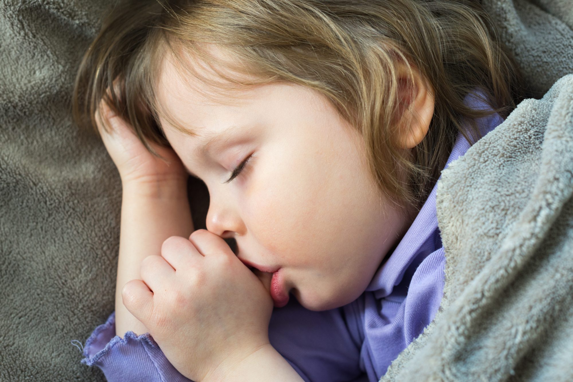 little girl laying under grey blanket sucking her thumb